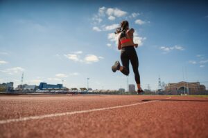 From below back view of teen girl in sportswear sprinting on running track at stadium under blue sky on summer day