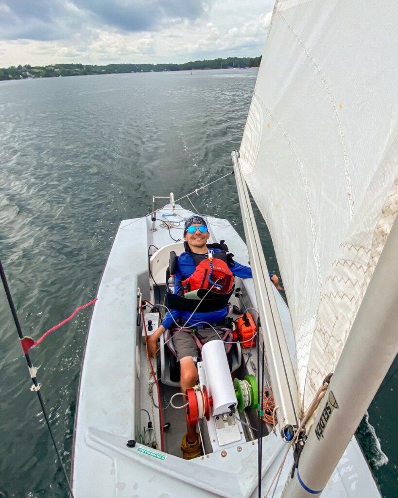 Un petit voilier vu à vol d'oiseau, avec le cockpit, le mât et la voile visibles. Un homme tétraplégique (Kevin Penny) est assis face à l'avant dans un siège personnalisé avec des sangles pour maintenir son torse. Il est entouré d'équipements d'assistance : un guindeau avec des cordes enroulées autour des bobines, une boîte avec un joystick, un boîtier orange contenant une batterie et un jeu de pailles placé sous son menton. Il porte une chemise à manches longues bleu roi, des lunettes à miroir et sourit à la caméra.